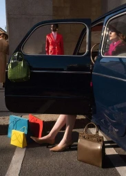 Two women posing around a dark blue vintage car with colorful handbags placed on the car door and pavement.