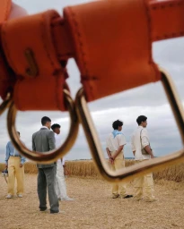 Close-up of an orange leather belt buckle framing several men standing in a dry grassy field under an overcast sky.