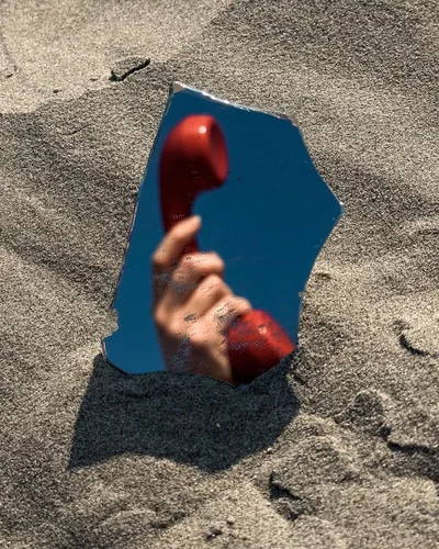 A hand holding a red telephone handset reflected in a broken mirror embedded in sand.