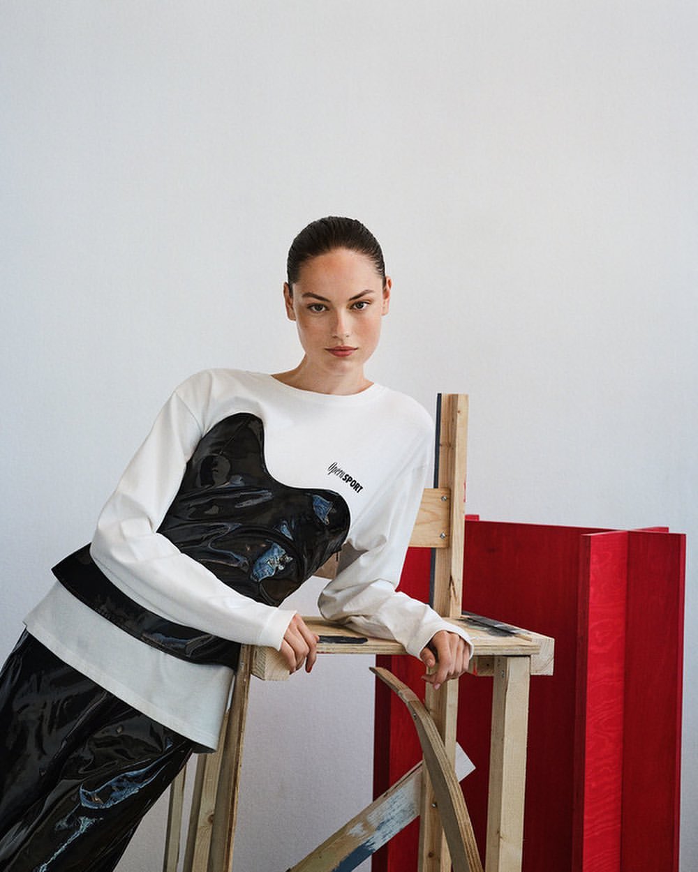 Fashionable woman in a white long-sleeve shirt with black print, shiny black pants, and a wooden chair, posing in a studio setting.