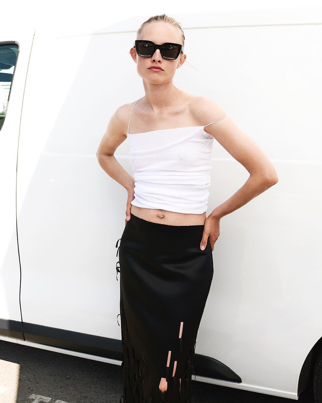 Woman poses in front of white van wearing white top, black skirt, and sunglasses, showcasing fashion and street style.