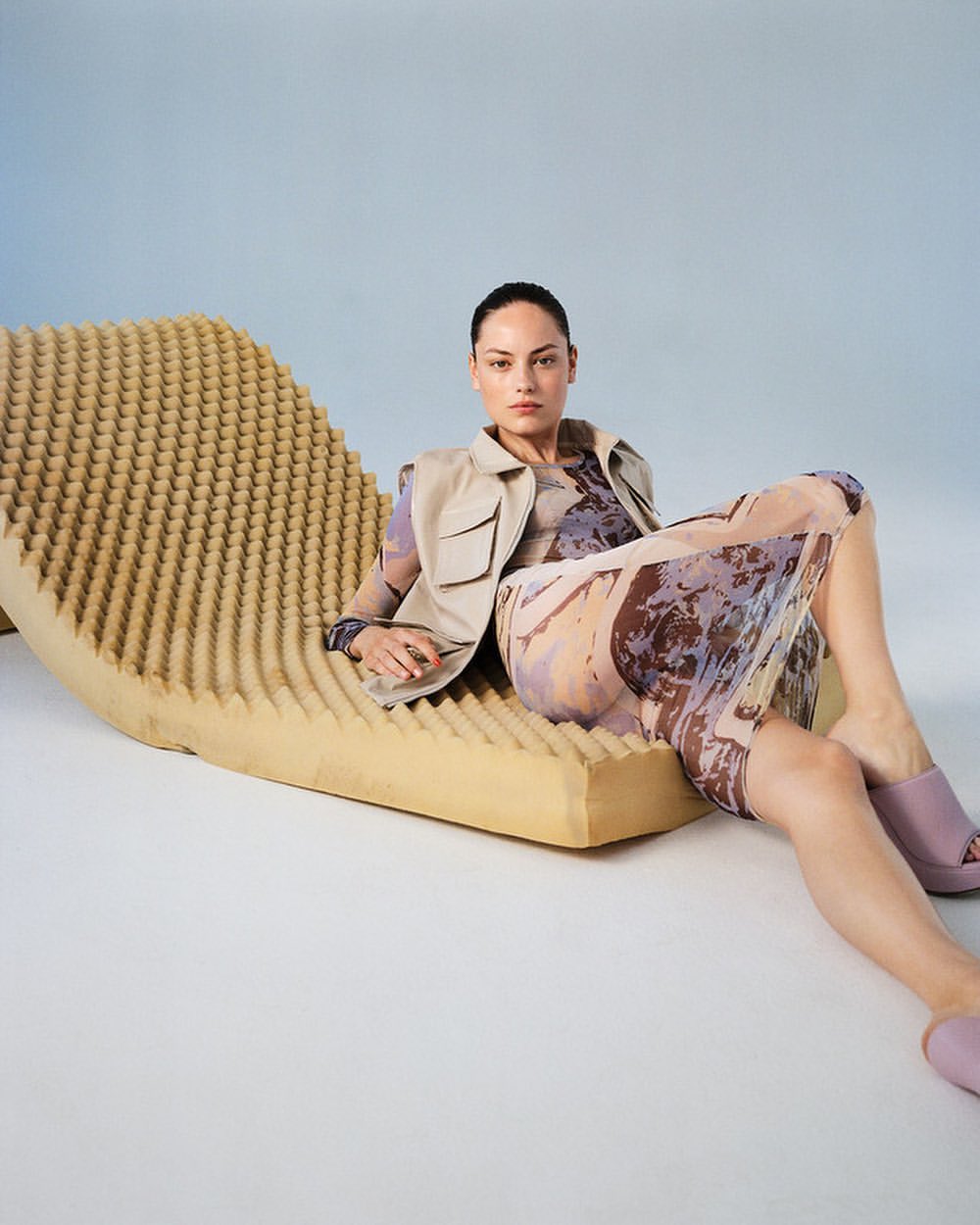 Adult woman poses on a textured foam lounger in a light-filled studio wearing a jacket, printed dress, and lilac platform shoes.