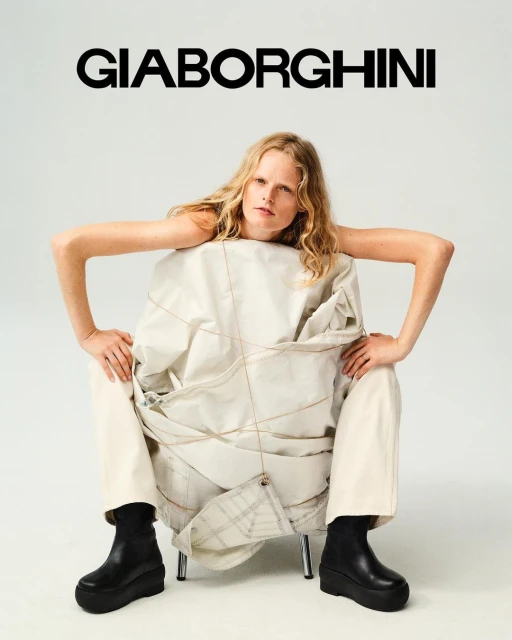 A fair-skinned adult woman with wavy blonde hair sits in a chair wrapped in beige fabric wearing black platform boots in a studio.