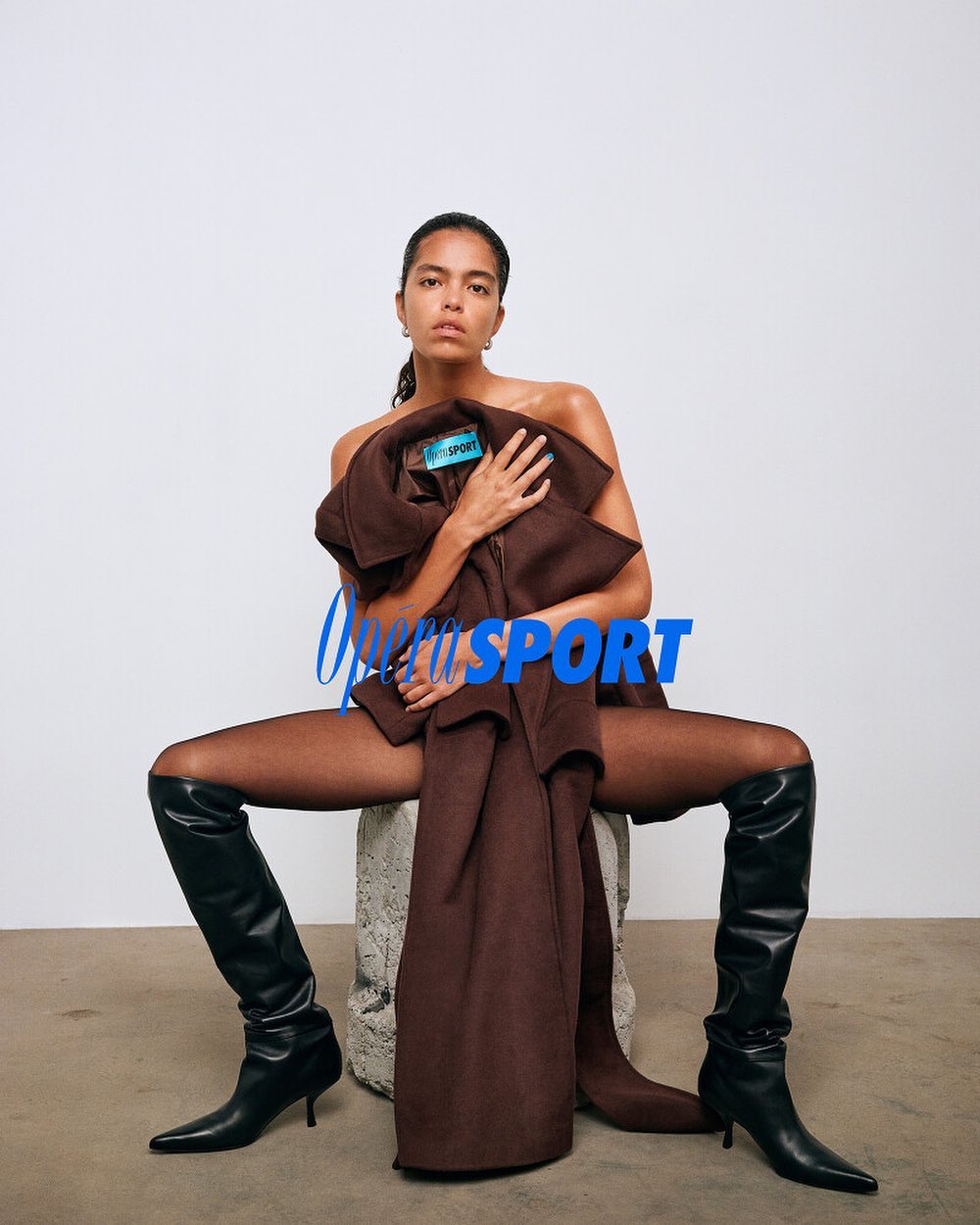 A woman poses in black boots and brown clothing, sitting on a textured gray pillar against a plain white backdrop in a studio setting.