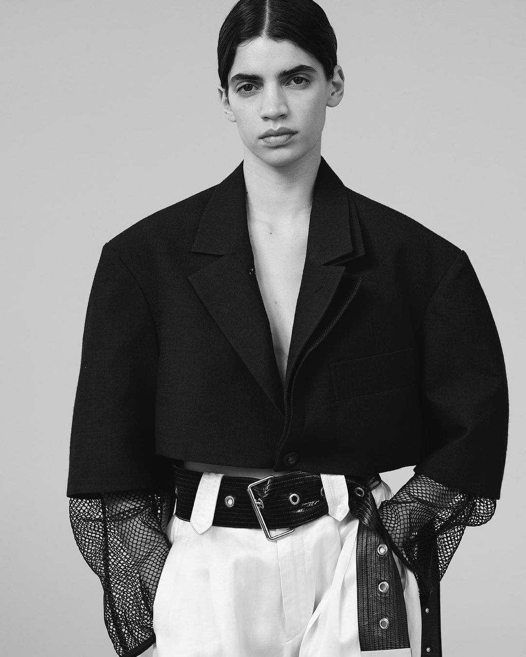Black-and-white fashion shot of a young adult woman wearing an oversized blazer and white pants with black belt in a studio setting.