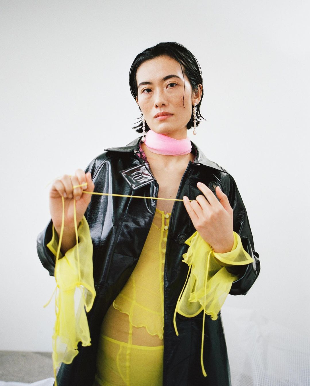 Woman in see-through yellow top and vinyl coat poses indoors with an elastic band, set against a bright white backdrop.