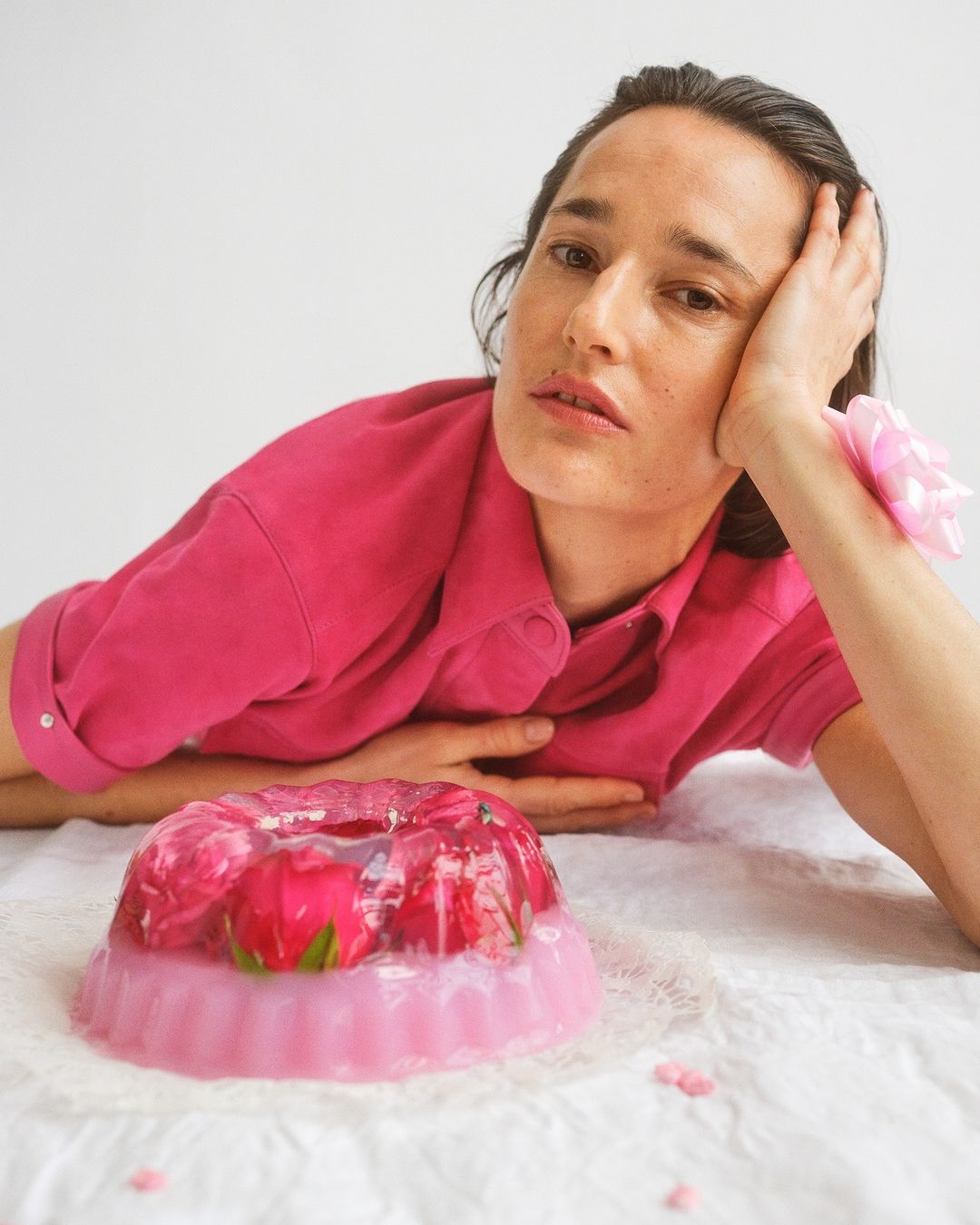 Adult female poses with a pink jelly dessert with roses on a white background wearing a hot pink shirt in a studio setting.