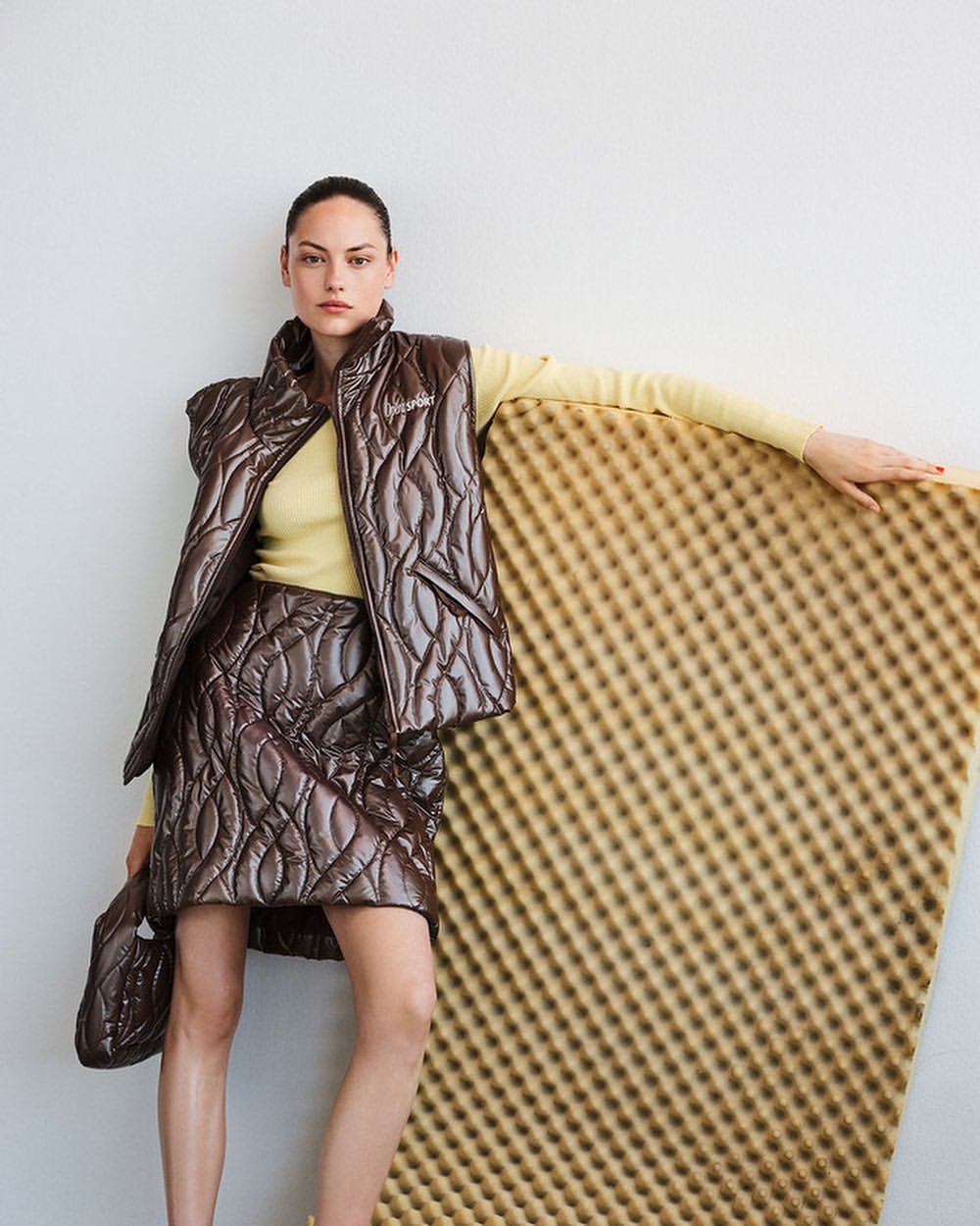 Woman poses in a brown quilted vest and skirt with a yellow top, leaning on a textured foam board in an indoor studio setting.