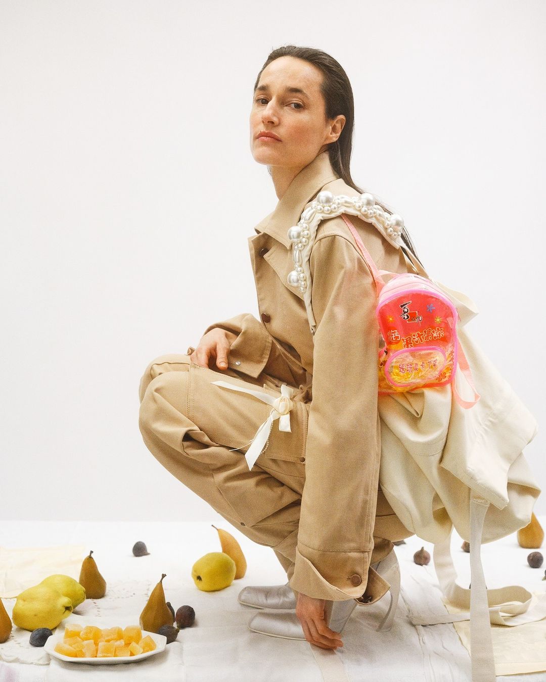Woman in trench coat and fashion backpack crouches in studio with fruit still life on white backdrop in a studio setting.