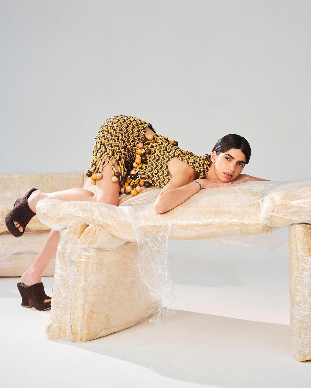A woman lies on a clear plastic-wrapped couch wearing a knit dress with pom poms and platform shoes in a neutral studio setting.