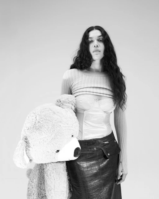 Monochrome portrait of a person with long dark hair, a light-colored top, and a dark skirt posing with a large teddy bear.