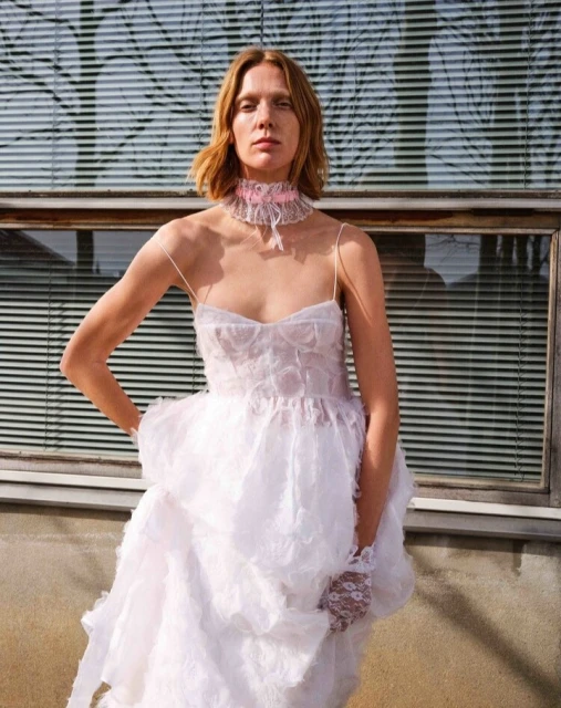 Woman in white layered dress with lace gloves and choker poses against building with blinds and neutral light in an outdoor setting.