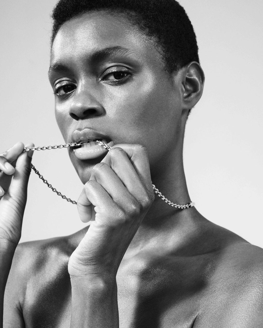 Close-up black and white studio portrait of a bare-shouldered African woman holding a silver chain necklace in her hands and mouth.