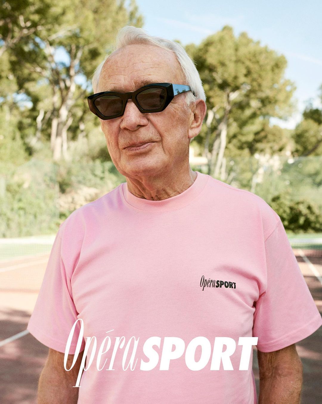 An elderly man wearing sunglasses and a pink T-shirt stands outside on a sunny day with trees in the background. The Opera Sport logo is visible.