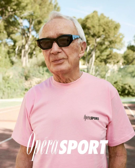 An elderly man wearing sunglasses and a pink T-shirt stands outside on a sunny day with trees in the background. The Opera Sport logo is visible.