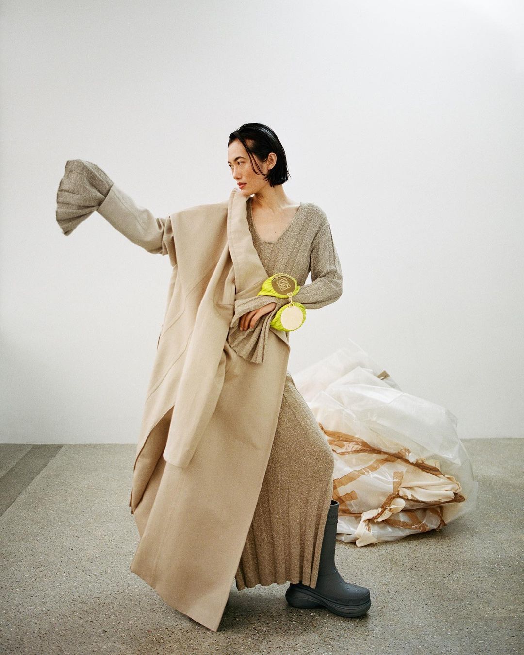 Fashion shot of a woman in neutral knit dress and coat with gray boots posing in a photo studio with a white plastic bag.