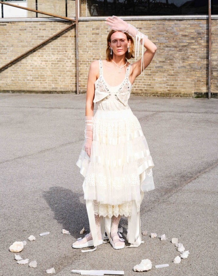 A stylish adult woman in a formal dress strikes a pose in an urban environment with a brick wall in the background on a sunny day.