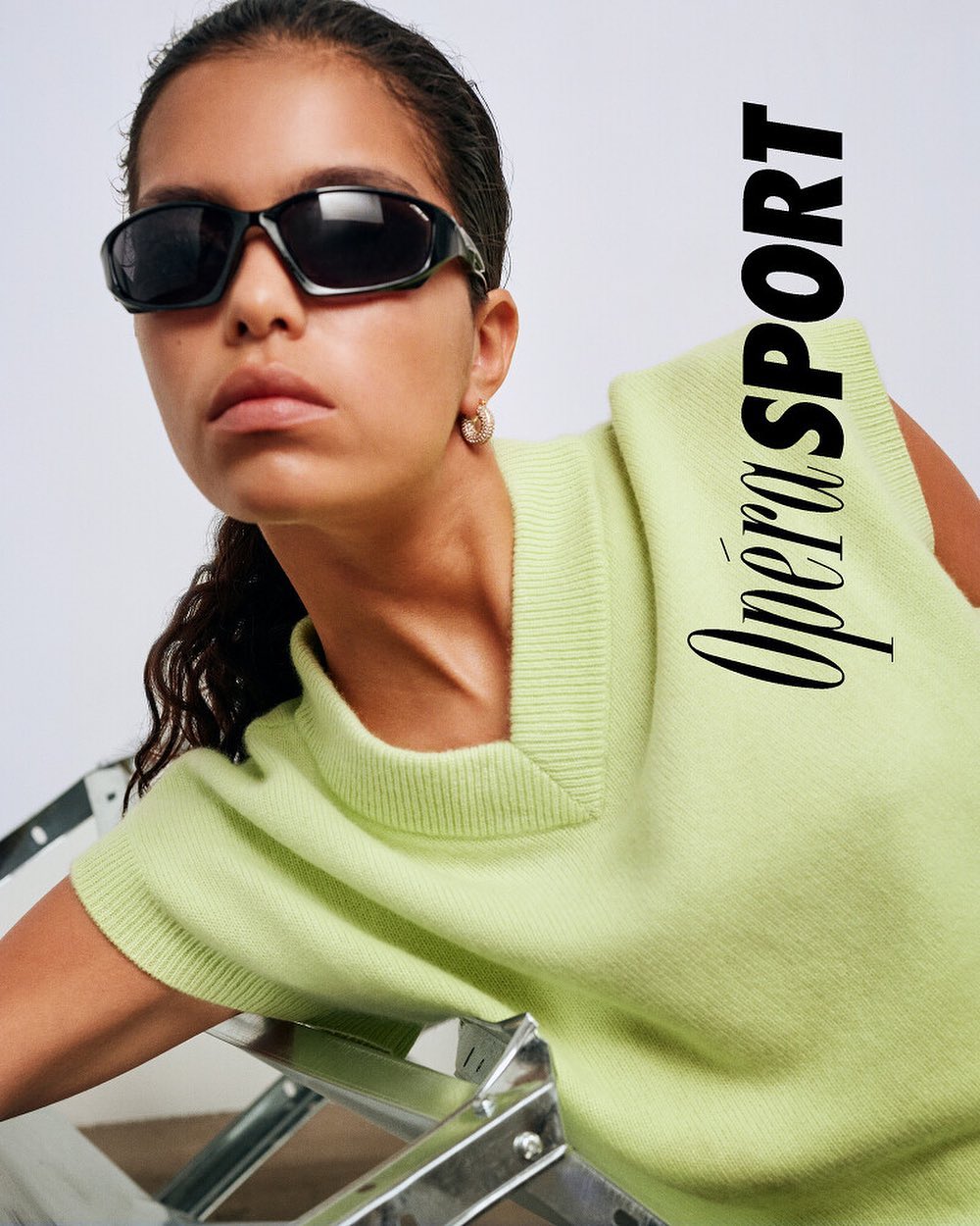 Portrait of a young adult woman wearing sunglasses and a bright green top, posing against a light background with metal chair.