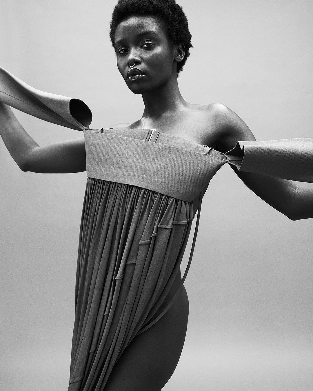 Black and white portrait of a Black woman in a draped dress with a nose ring, posing against a plain gray backdrop in a photo studio.