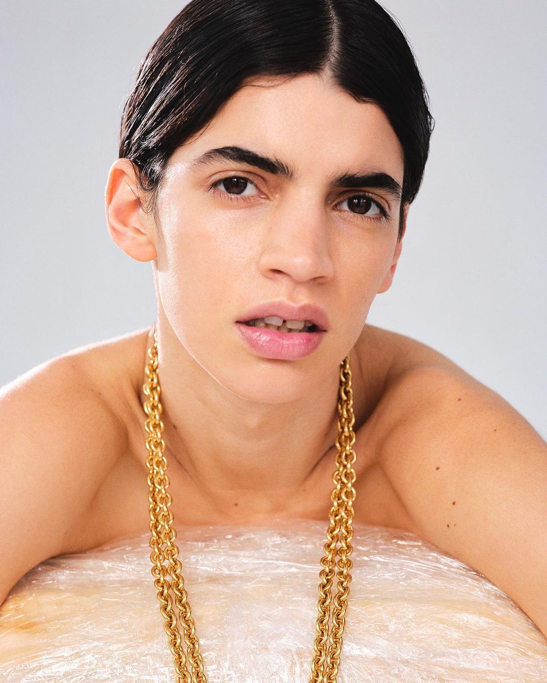 Close-up portrait of a young adult with dark hair wearing a thick gold chain necklace, posing in front of a light gray background.