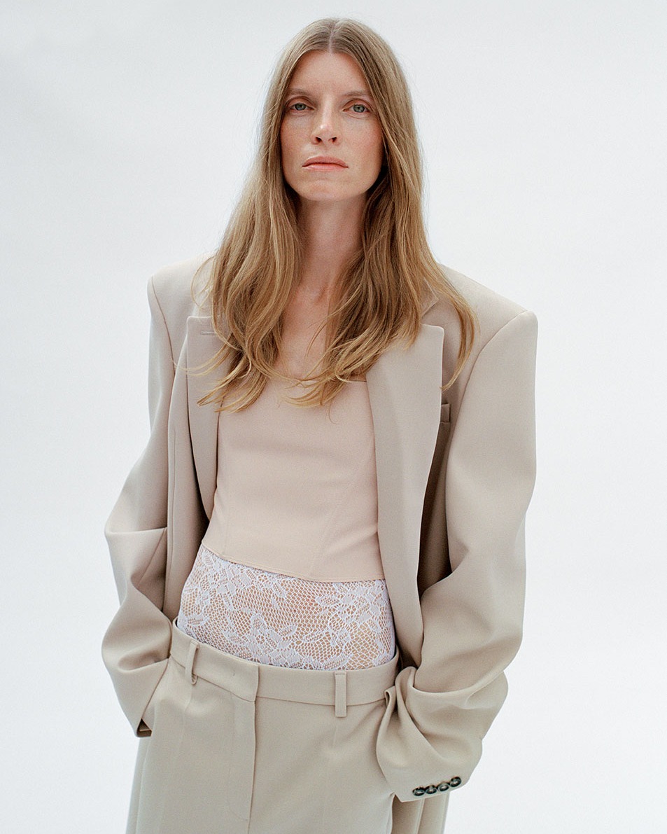 A fair-skinned woman with long blonde hair poses in a beige blazer, top, and slacks against a white backdrop in a studio setting.