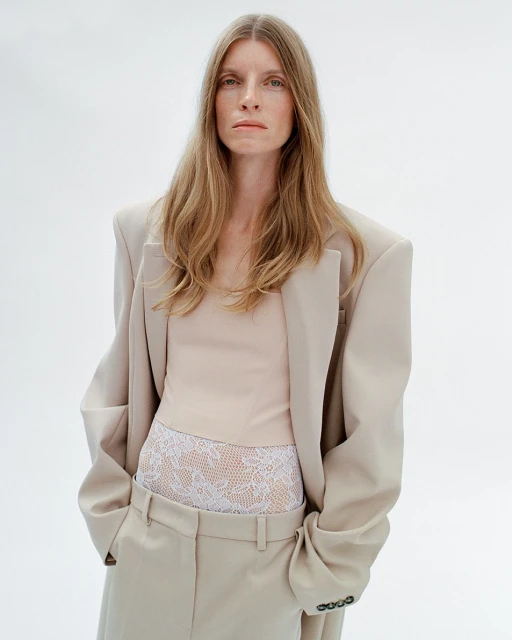 A fair-skinned woman with long blonde hair poses in a beige blazer, top, and slacks against a white backdrop in a studio setting.
