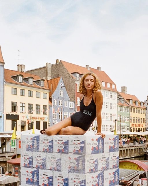Young woman in swimsuit sitting on stacked beverage cases in urban Copenhagen, Denmark, with colorful buildings and canal view.