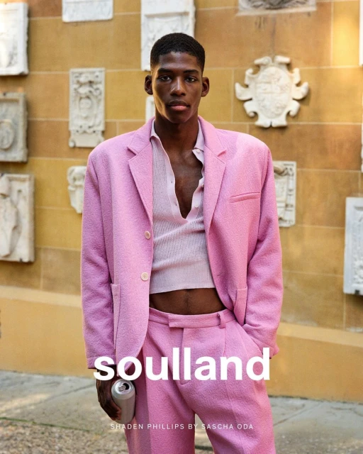 A male model in a pink suit poses outdoors with a drink, showcasing a fashion editorial against a textured wall. The photograph emphasizes style.