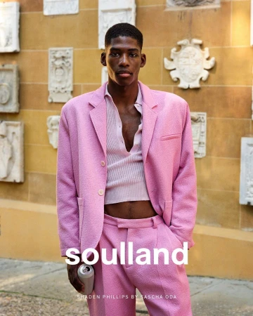 A male model in a pink suit poses outdoors with a drink, showcasing a fashion editorial against a textured wall. The photograph emphasizes style.