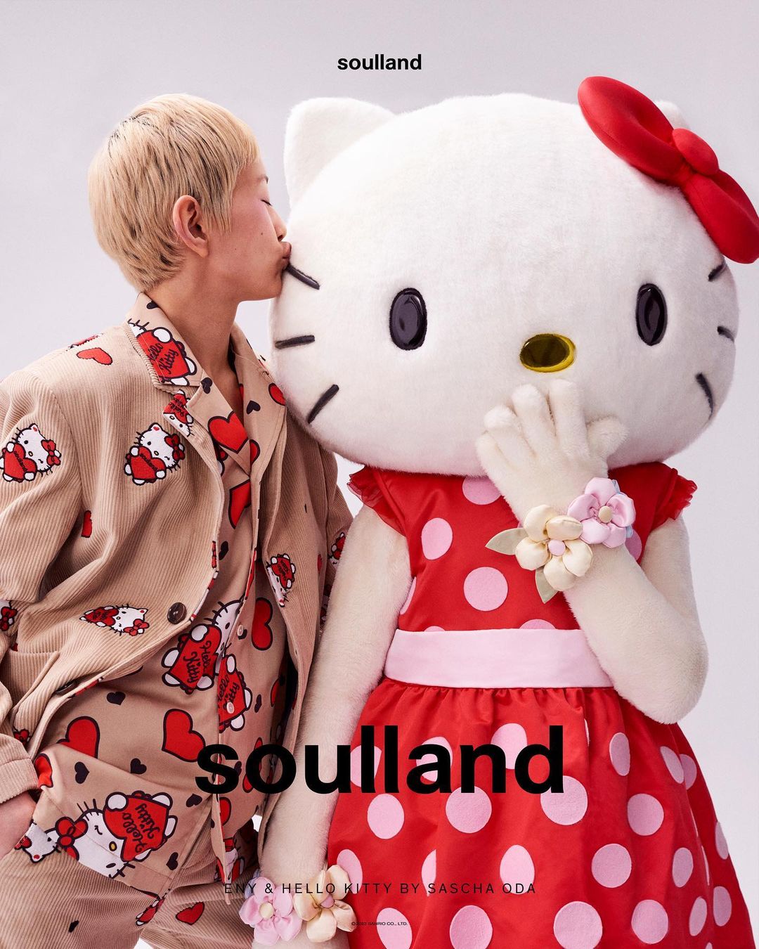 Asian adult wearing patterned pajamas kisses a Hello Kitty mascot wearing a polka dot dress in a bright photo studio setting.