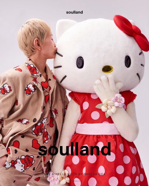 Asian adult wearing patterned pajamas kisses a Hello Kitty mascot wearing a polka dot dress in a bright photo studio setting.