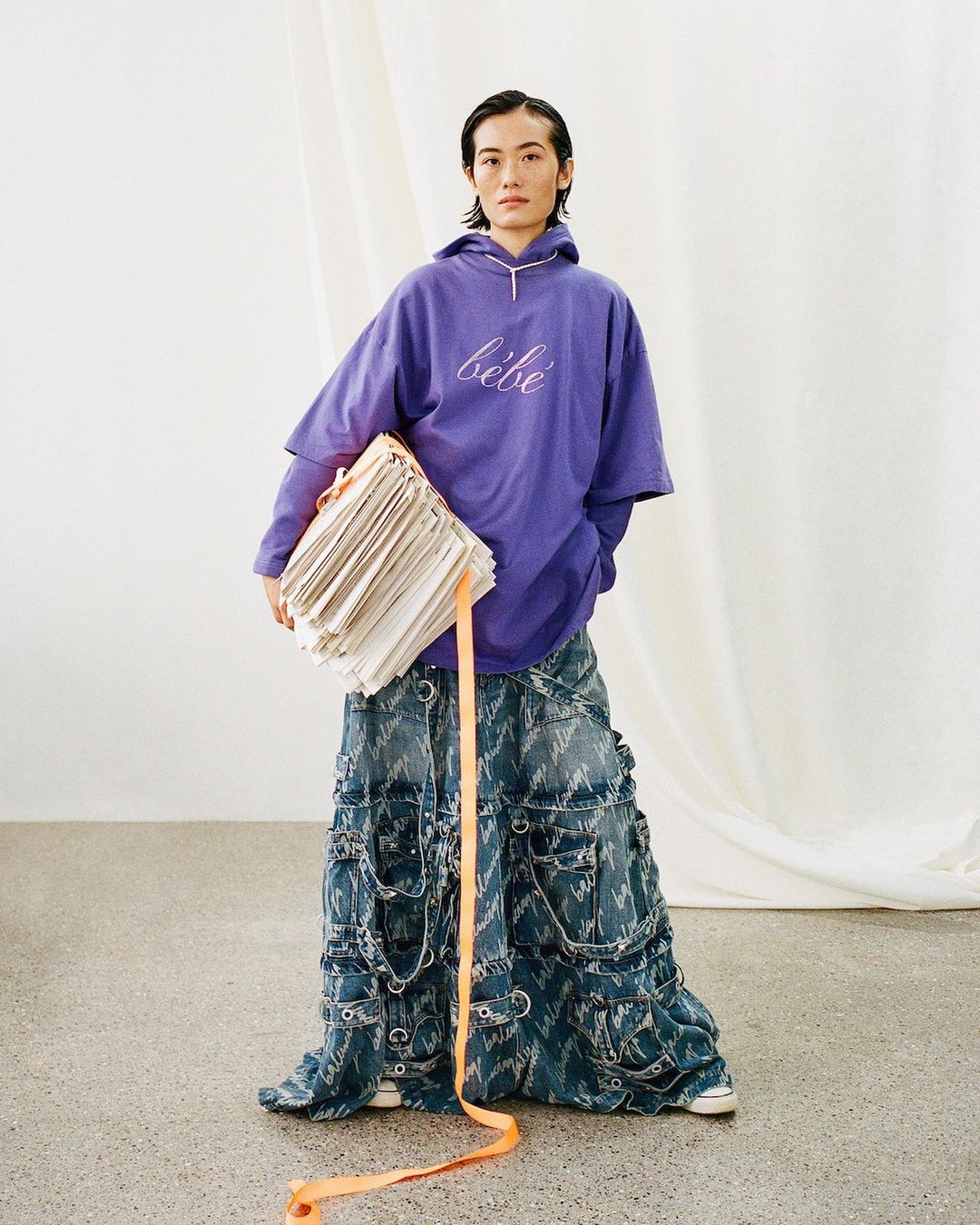 Non-binary person in a purple 'bebe' hoodie and patchwork jeans stands in a bright studio holding a stack of papers.