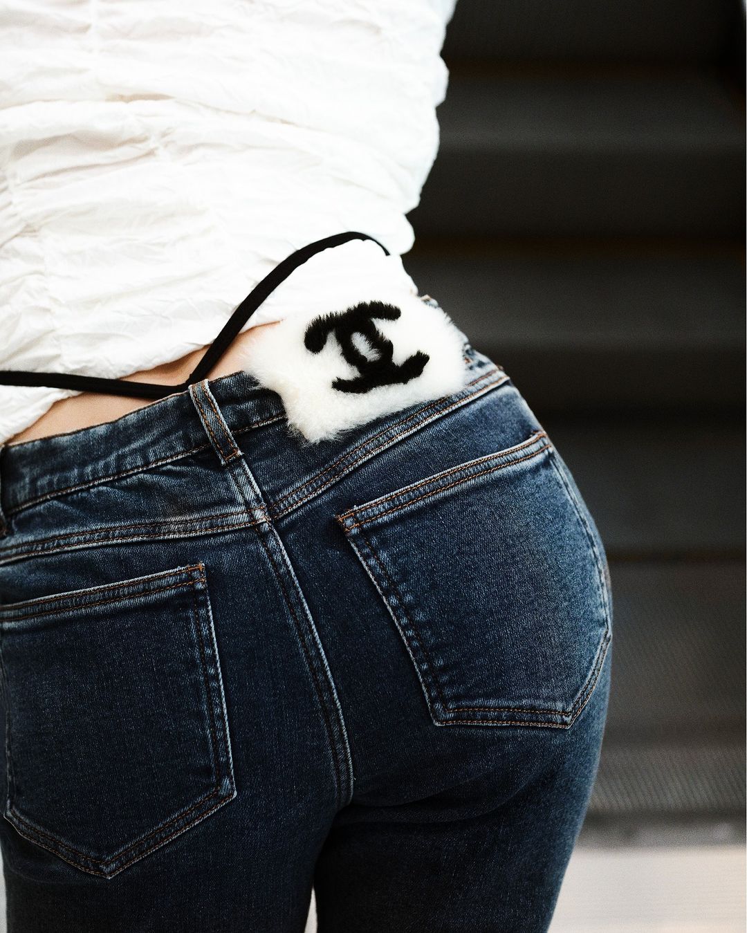 Close-up of a woman wearing dark denim jeans with a white shirt. A designer accessory with a recognizable logo is visible.