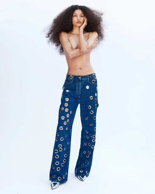 Fashion shot of a young adult woman posing in blue denim pants with silver metal eyelets and pointed silver heels on a white background.