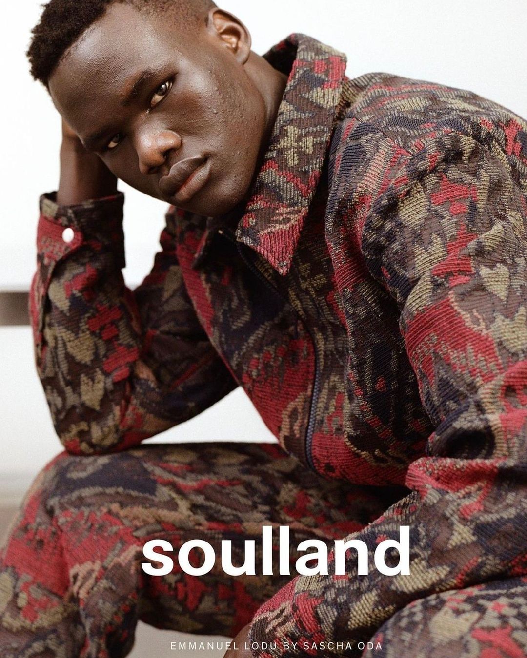 A dark-skinned man is featured in a medium shot, wearing a patterned jacket and pants while posing indoors with a neutral expression.