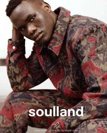 A dark-skinned man is featured in a medium shot, wearing a patterned jacket and pants while posing indoors with a neutral expression.