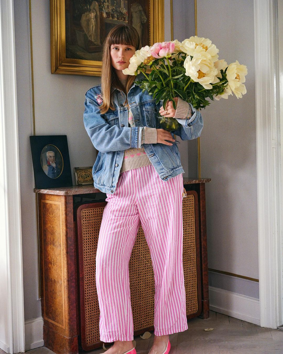 Young woman holding large bouquet of white and pink peonies indoors. She wears a denim jacket and pink striped trousers in bright daylight.