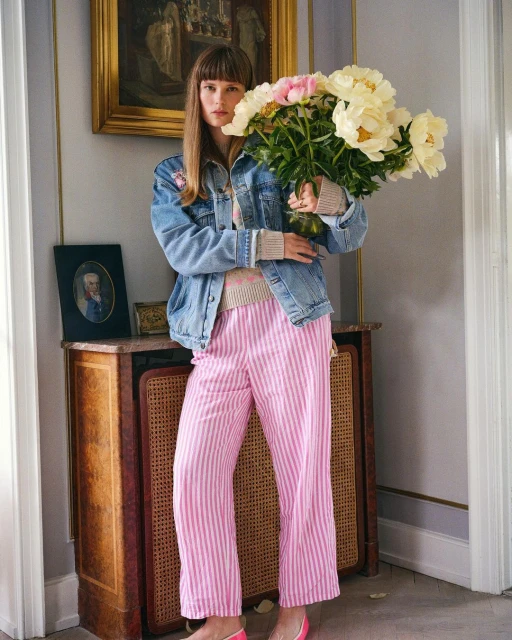Young woman holding large bouquet of white and pink peonies indoors. She wears a denim jacket and pink striped trousers in bright daylight.