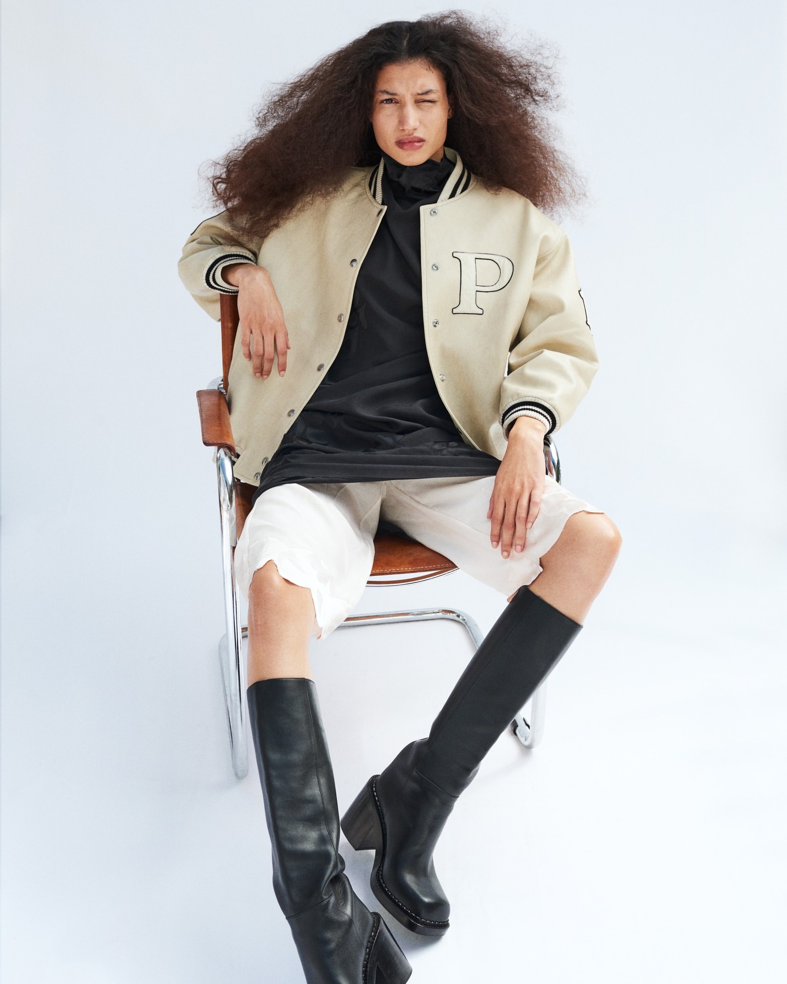 Fashion portrait of an adult woman with big hair wearing a beige jacket, black shirt, white shorts, and black boots, seated in a studio.