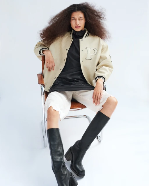 Fashion portrait of an adult woman with big hair wearing a beige jacket, black shirt, white shorts, and black boots, seated in a studio.