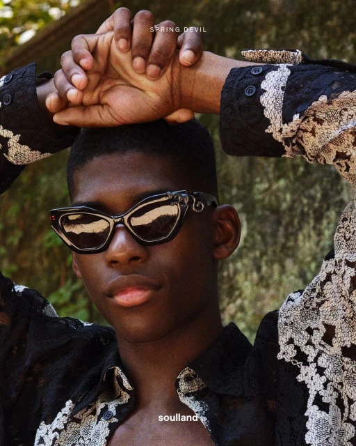 Close-up studio portrait of a Black man with short hair wearing stylish sunglasses and a lacy black shirt against a blurred background.