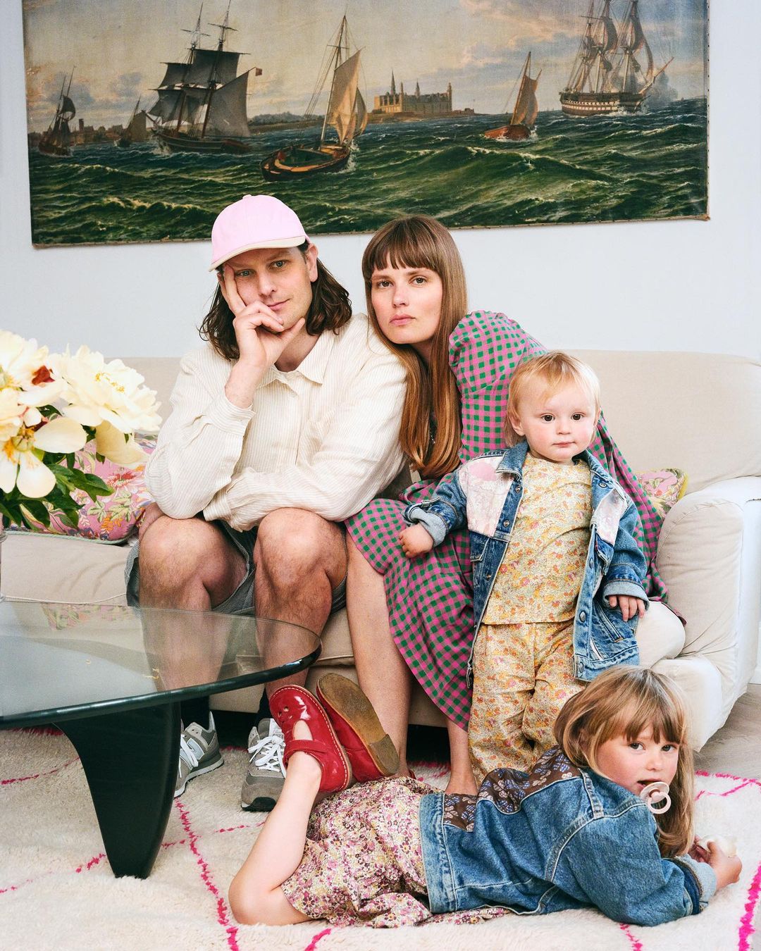 Family portrait featuring a man, a woman, and two young children posing indoors in front of a tapestry depicting sailing ships.