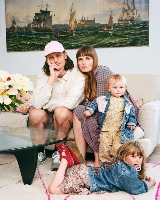 Family portrait featuring a man, a woman, and two young children posing indoors in front of a tapestry depicting sailing ships.