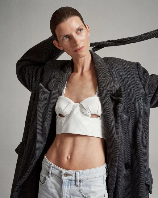 A stylish adult woman poses in a photo studio, wearing a gray blazer, white top, and jeans against a neutral backdrop.