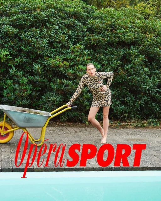 Woman in leopard dress poses with a wheelbarrow by pool, against hedge backdrop. Fashion editorial, outdoor, casual. 