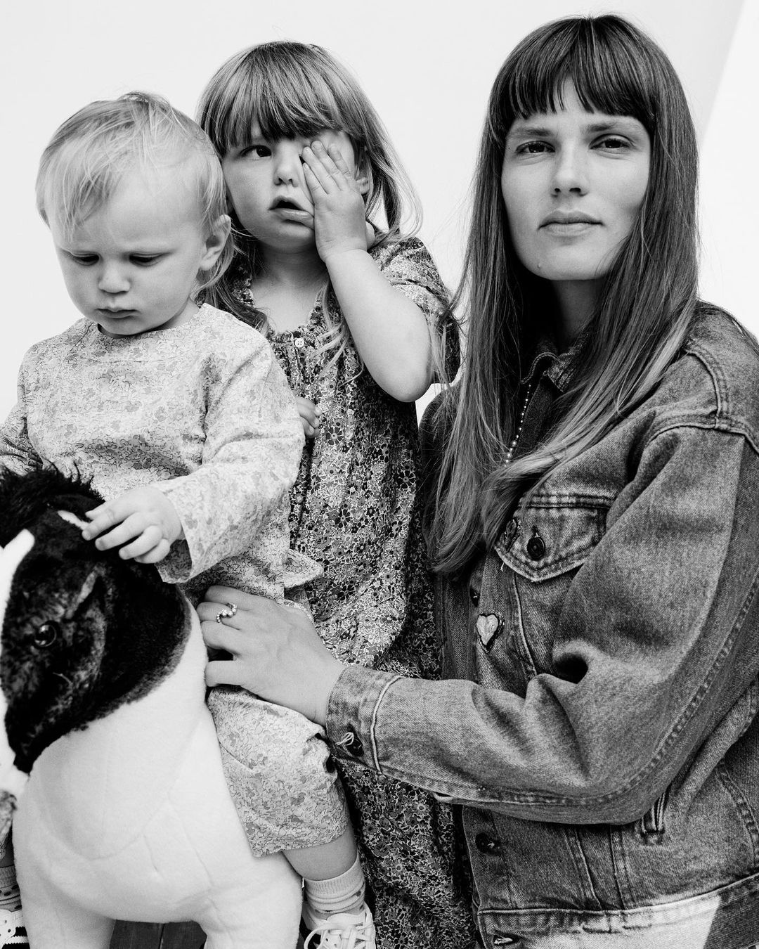Black and white portrait of a woman and two young children, one seated on a toy horse, in a family or lifestyle setting, shot in studio.