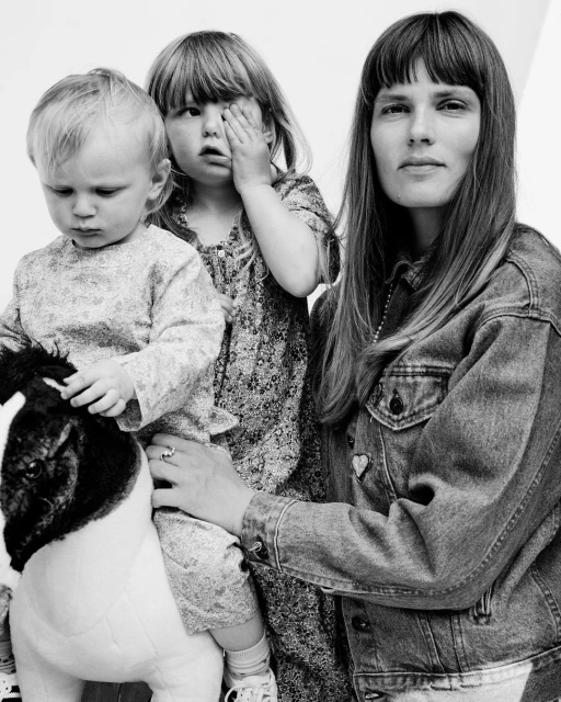 Black and white portrait of a woman and two young children, one seated on a toy horse, in a family or lifestyle setting, shot in studio.