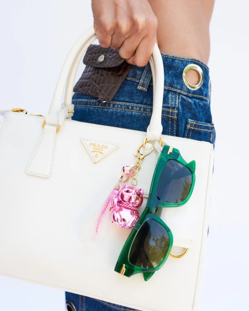 Close-up of a person holding a white Prada handbag with green sunglasses and a pink charm, showcasing fashion accessories.