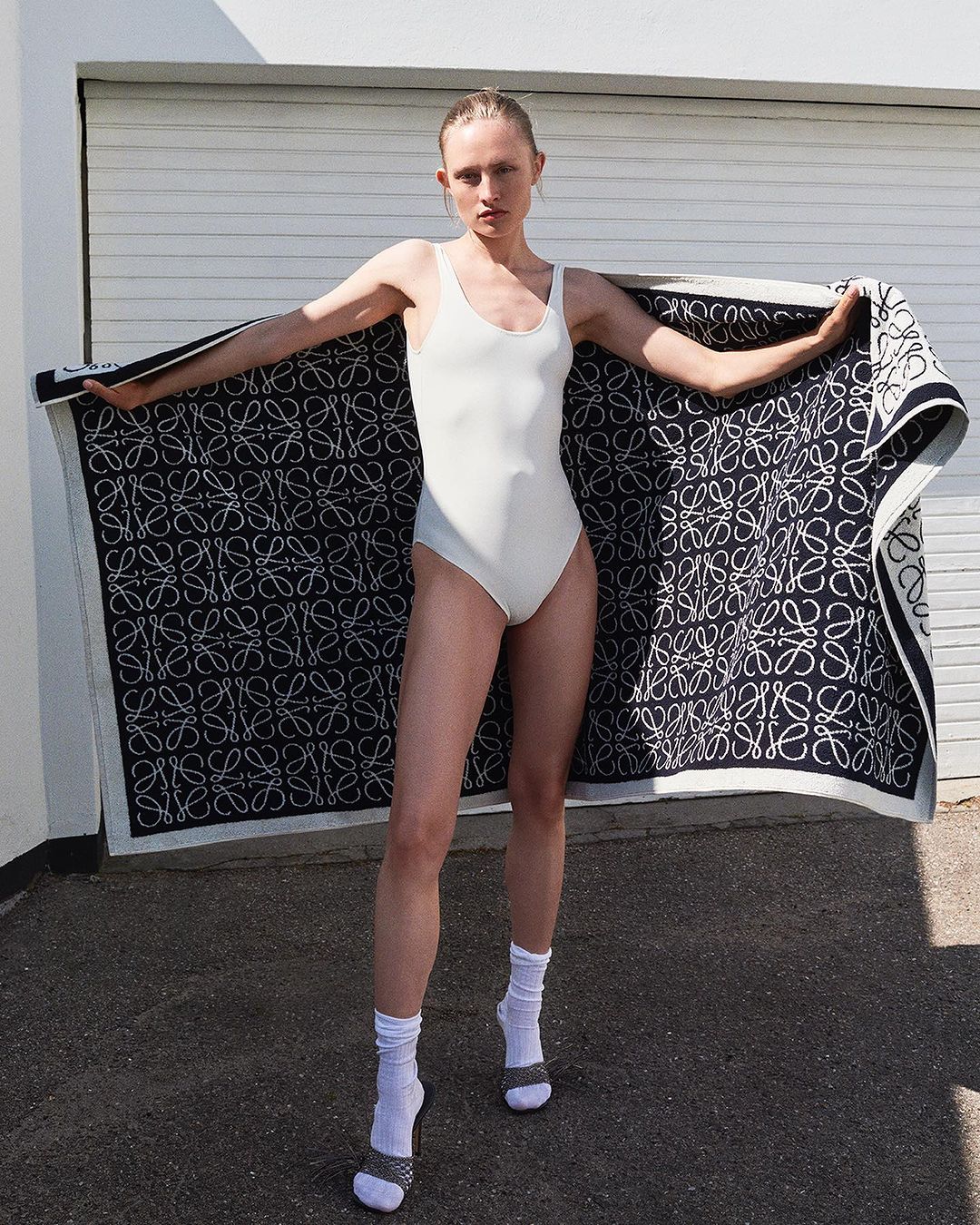 Fashion shot of a woman in a white bathing suit holding a large patterned Loewe towel outdoors in front of a white wall.