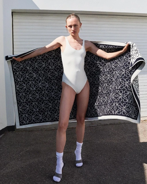 Fashion shot of a woman in a white bathing suit holding a large patterned Loewe towel outdoors in front of a white wall.
